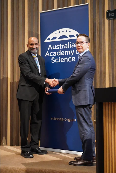 A person receiving a certificate in a cylinder from another person. They stand shaking hands in front of a banner with the logo of the Australian Academy of Science. 