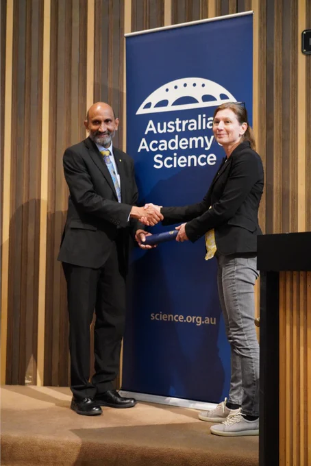 A person receiving a certificate in a cylinder from another person. They stand shaking hands in front of a banner with the logo of the Australian Academy of Science. 