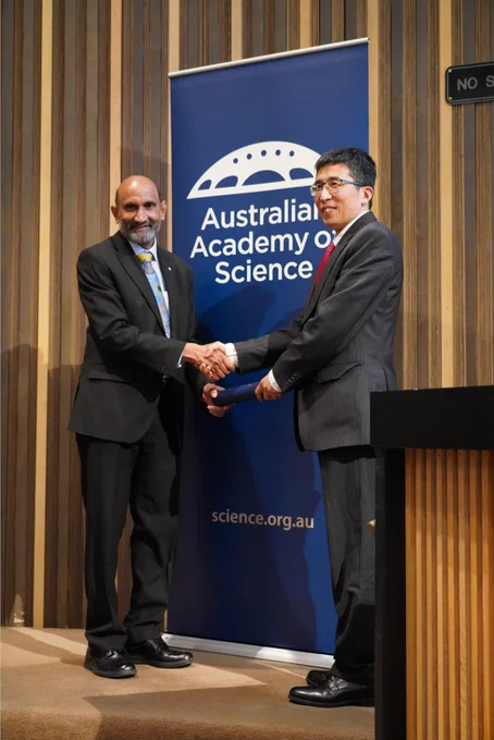 A person receiving a certificate in a cylinder from another person. They stand shaking hands in front of a banner with the logo of the Australian Academy of Science. 