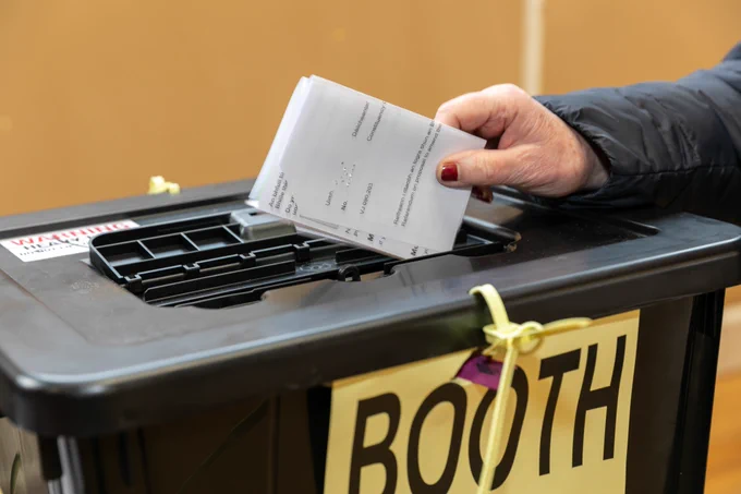 A hand places a ballot paper into the black ballot box. 
