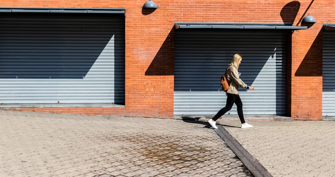 A blonde woman in black jeans and a khaki jacket walks past a series of garage doors.