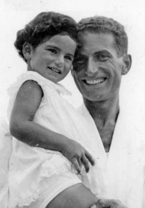 Black and white photo of a smiling man holding a young child, both dressed in light clothing.
