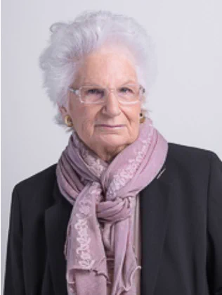 Portrait of an elderly woman with white hair, wearing a black blazer and a light pink scarf, set against a plain light background.
