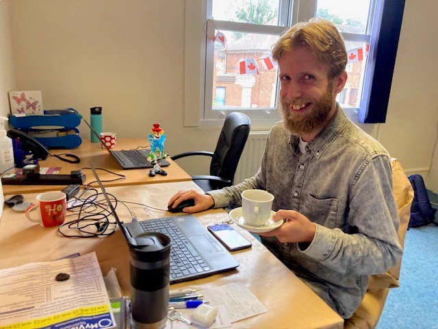 Image description: A photograph of a man sat at a desk. He is looking at the camera and smiling, and is holding a cup and saucer in one hand. There are several Canadian flags decorating the window behind him.
