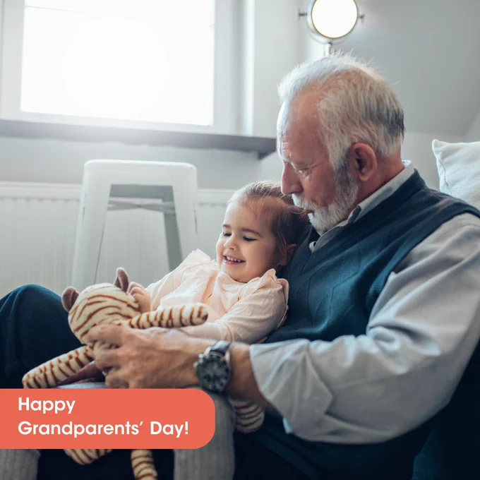 An image of a man sitting on a chair in a living room. On his lap is a child holding a stuffed animal. 