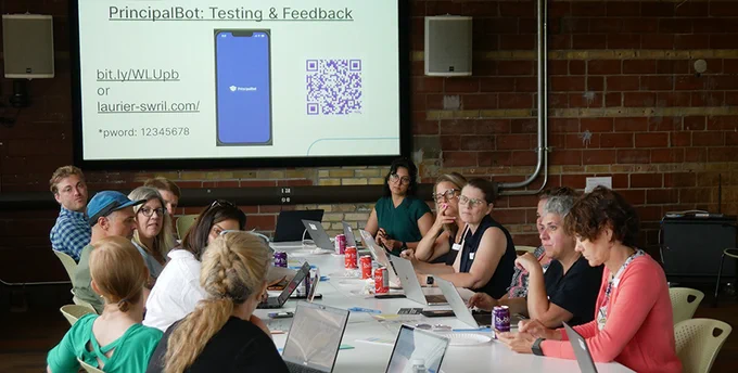 A group of about a dozen adults sitting around a long table in a boardroom. Projected on the screen is the Principal BOT, the AI Chatbot the principals are testing. They are smiling and appear to be discussing the tool and what is projected on the screen.