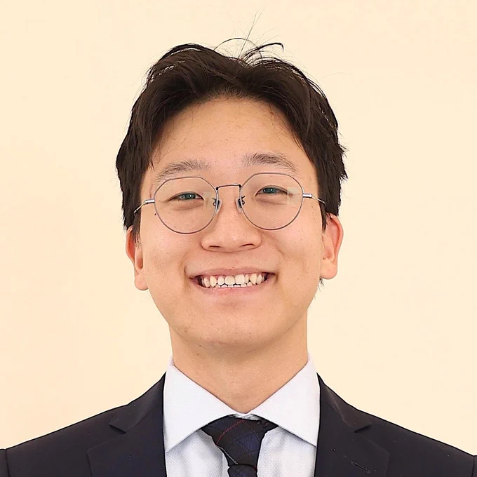 Dr. Alex Bak smiles against a pale yellow background. He is a wearing a black suit and tie. 
