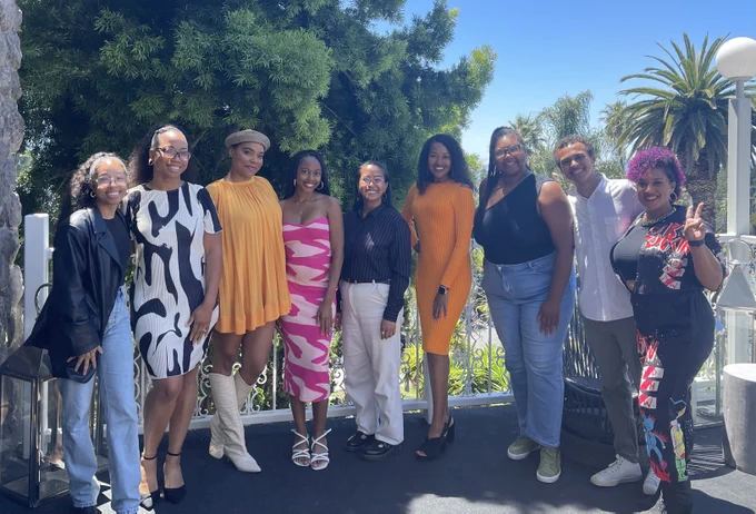 A group of young BIPOC adults dressed in business casual attire, standing close to each other and on a patio against a lush green background of leafy trees and palms.