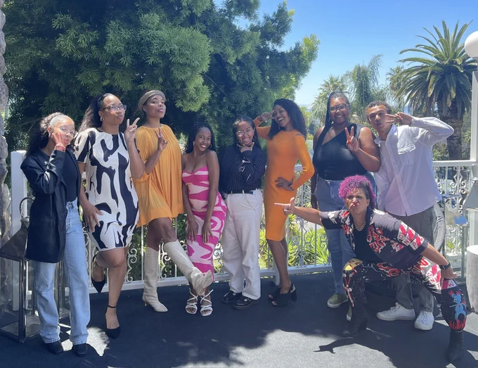 A group of young BIPOC adults dressed in business casual attire, standing close to each other and on a patio against a lush green background of leafy trees and palms. In this picture, they're making energetic, silly faces and poses (including peace signs and standing in profile).