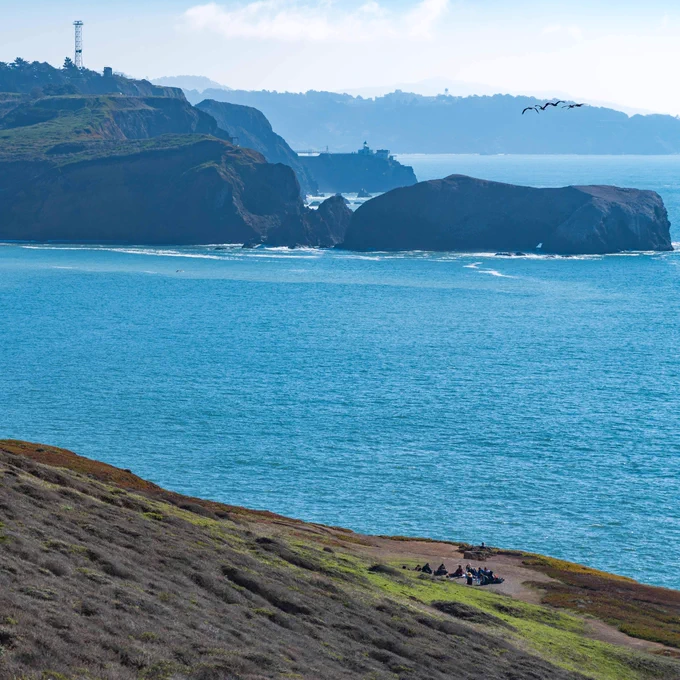 View from a scenic overlook looking over a brown hillside sloping down to the blue ocean, with large rock formations and the city of San Francisco in the distance