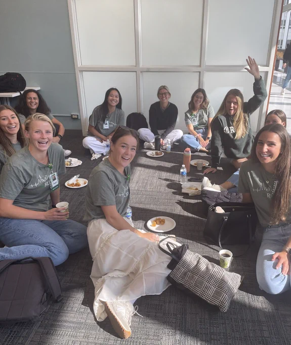 Group of first-year Rush Medical College students sitting on the floor and smiling while enjoying a meal together. Some are wearing green t-shirts with the Rush logo.