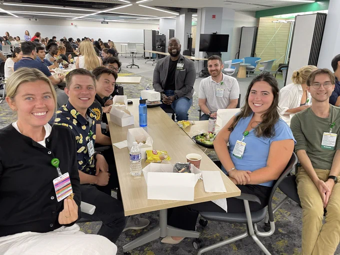 Group of first-year Rush Medical College students sitting at tables, enjoying a meal together. Everyone is smiling and appears engaged in conversation.