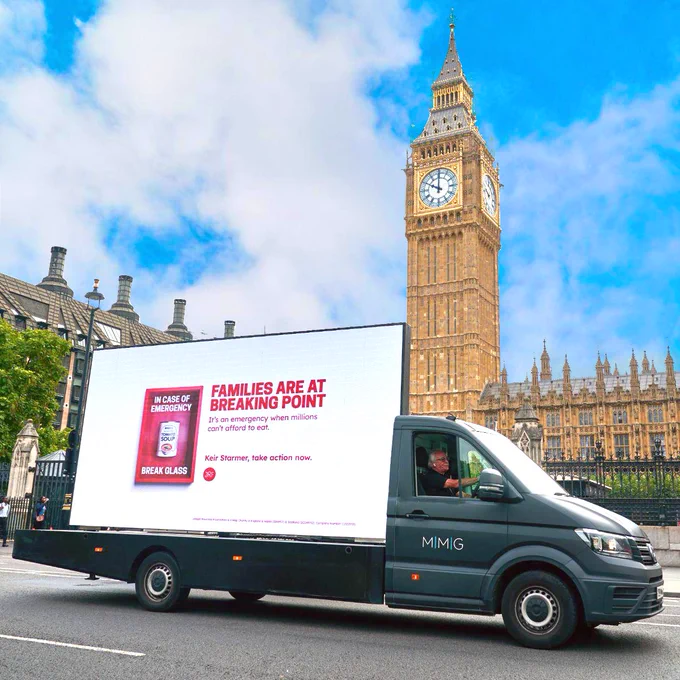 A 'digivan' driving past Elizabeth Tower at the Palace of Westminster. The image on the van includes an 'in case of emergency break glass' with a tin of soup inside. The text on the image says: 'Families are at breaking point. It's an emergency when millions can't afford to eat. Keir Starmer, take action now.'
