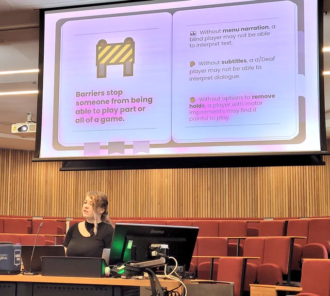 Cari mid-speaking in a lecture theatre with a screen above illustrating some of the barriers that stop playing games - including lack of menu narration, subtitles, and holds