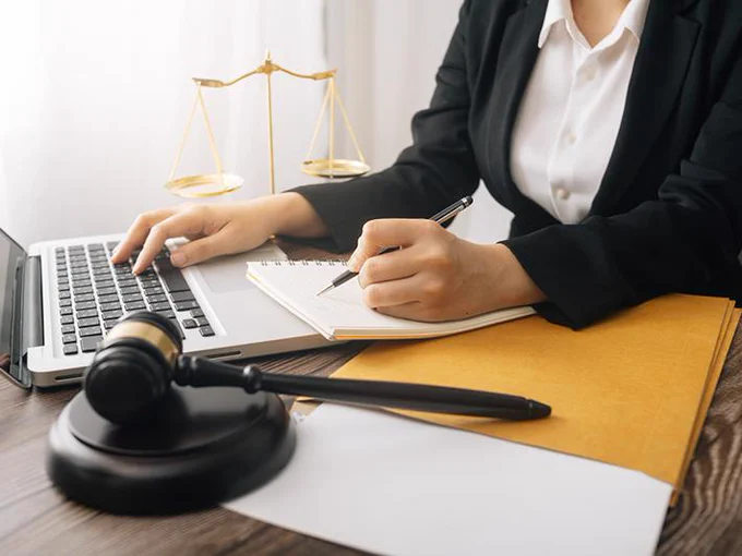 A woman whose face we can't see sits at a desk using a laptop and making notes