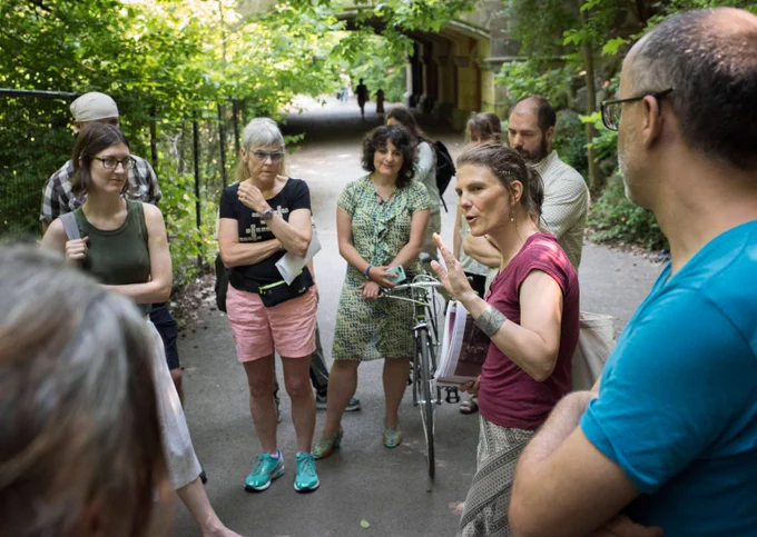 A member of the Turnstile Tours team guiding a small tour group of nine people through Prospect Park. The group is stopped on a paved path through the park woodlands as the tour guide carefully explains to the focused participants.