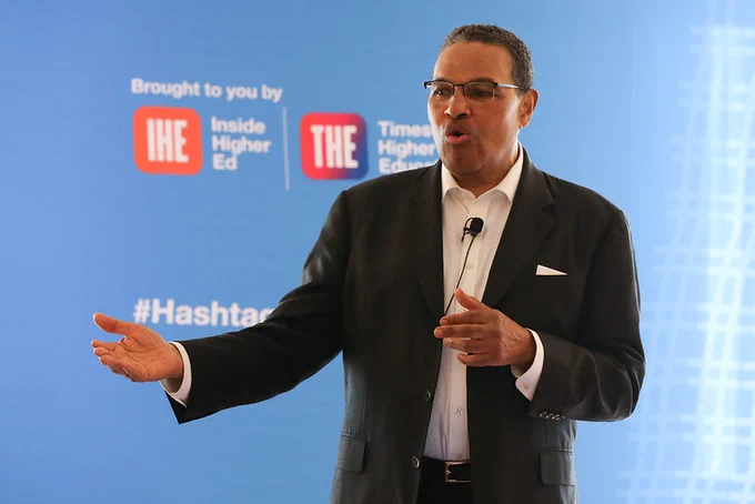 Speaker presenting at a conference with logos of Inside Higher Ed and  Times Higher Education in the background, alongside the hashtag.