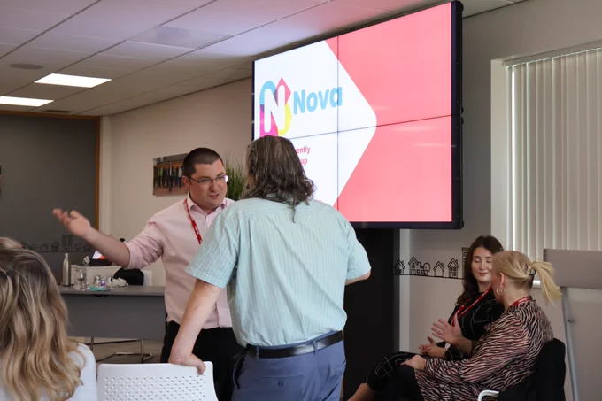 People stood and sat talking at the Thinking Differently Peer Meeting, there is a presentation screen with Nova's logo on.