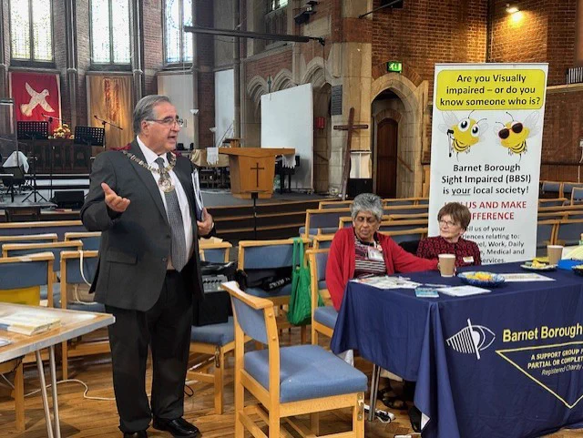 The Mayor of Barnet, Councillor Tony Vourou. speaking at the event. The Barnet Borough Sight Impaired stall and banner behind him.