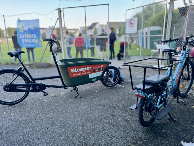 Two cargo bikes in the foreground - one green with cargo box at the front, the other blue with a cargo rack at the back. 

Blurred in the background is a sports pitch with parents watching.