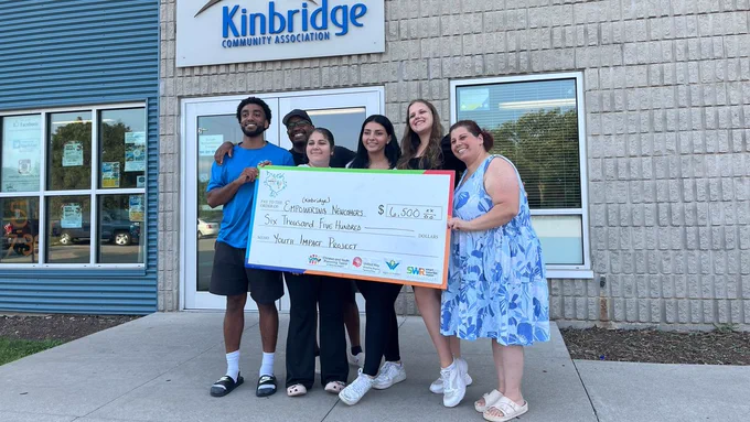 A group of youth and adults standing outside with a giant cardboard cheque. The cheque is written to Empowering Newcomers for $6,500. They are standing in front of a building that says "Kinbridge Community Association."
