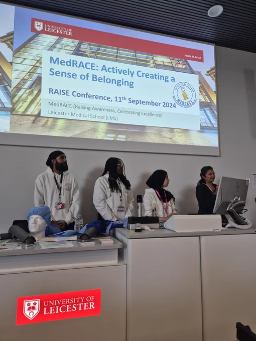 The MedRACE student co-chairs presenting in a University of Leicester lecture theatre. There are three students standing at the front of the lecture theatrem