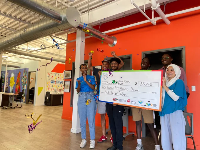A group of youth smiling, popping a party popper, and holding a giant cardboard cheque.