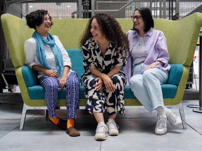 Three individuals seated on a sofa, laughing together in a modern, airy space. They are each dressed in casual, stylish attire.