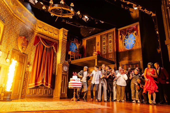 The West End cast of The Play That Goes Wrong on stage with original cast members on the production’s tenth anniversary with a birthday cake and a flame shooting out of the bin