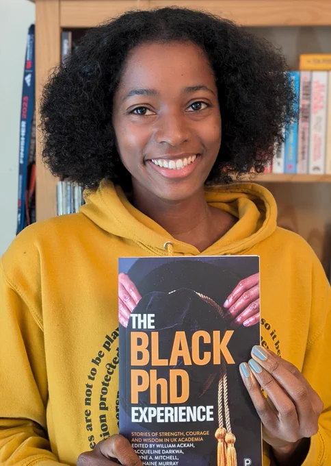 Woman wearing a yellow hoodie smiling in front of a bookcase, holding a paper back copy of 'The Black PhD Experience' book