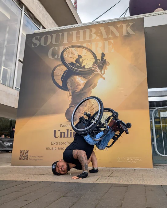 Nadenh Poan, a Cambodian male wheelchair dancer recreates the Lived Fiction and Unlimited poster image outside of the Southbank Centre. Behind Nadenh the huge poster depicts him in a gravity defying headstand. A haze of golden light spills around him as he throws his left arm forwards to the camera, balancing only on the side of his head and right arm, his lightweight black manual chair lifted above him. Photo by Chris Parkes.