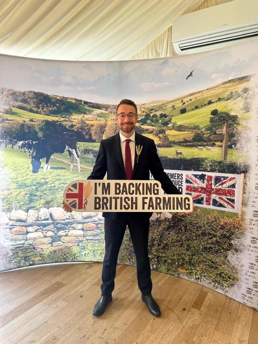 A photo of Luke holding a sign that says “I’m backing British farming” with a rural background