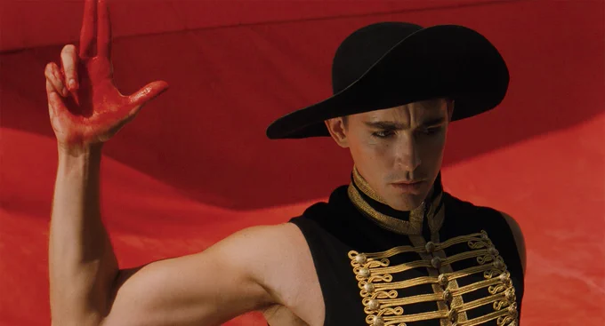 A person wearing a black hat and a black vest with gold detailing, holding their arm out to their side and making the shape of a gun with their red-stained hand. 