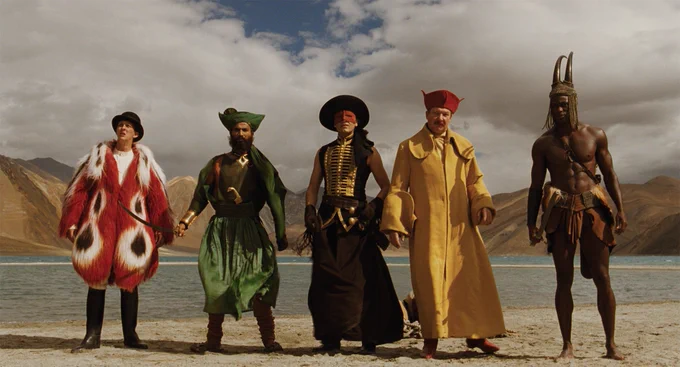 Five people wearing extravagent costumes, standing in front of a lake, with a mountainous terrain in the background