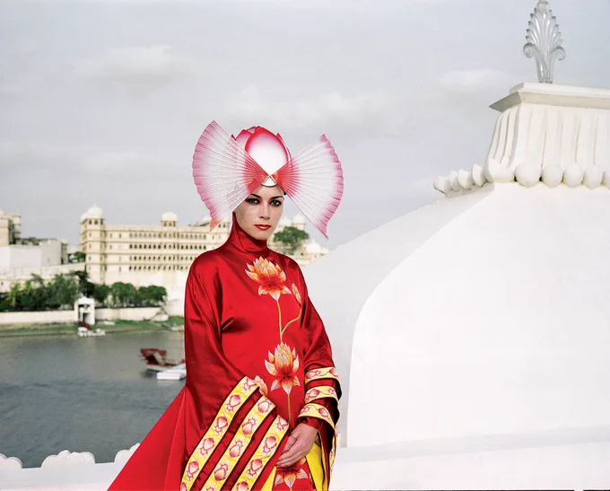 A person wearing an extravagent red cloak and headdress, standing by a large white building.