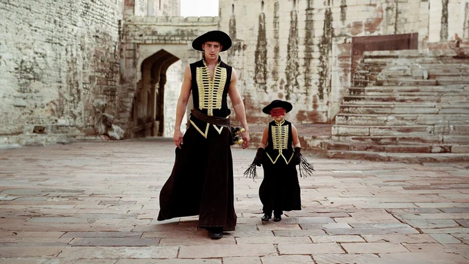 Two people, both wearing black hats, black vests with gold detailing and black trousers, walking side by side in a courtyard.