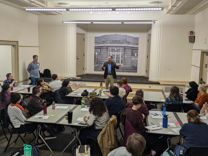Sec. Hobbs presenting to a group of people seated at tables in a classroom setting, with a large black and white photo of a building projected on the screen behind.