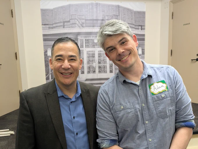 Sec. Hobbs and Adam Davis (Game to Grow)  smile in front of a banner that reads "Everett Public Library." One person is wearing a suit, and the other is in a denim shirt with a name tag labeled "Adam.