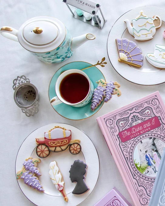 tea cups, tea pots, and Bridgerton-themed cookies (carriage, fan, feather, lilac) on a white tablecloth next to a pink hardcover special edition book of The Duke and I