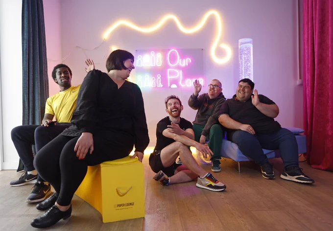 A group of five people smiling and waving in a brightly lit room with colorful neon lights. The sign in the background reads 'Our Place.' One person sits on a yellow block labeled 'Paper Lounge,' while others sit on or near a couch. The mood is joyful and relaxed."