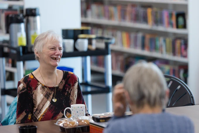 An older woman smiles as she sits at a table with a mug.