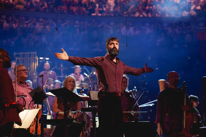 Jules Buckley stands in front of his Orchestra, facing the audience with his arms outstretched to accept rapturous applause. He wears a maroon shirt and dark trousers, and the ensemble wear difference shades of mauve and maroon, standing to face the audience. 