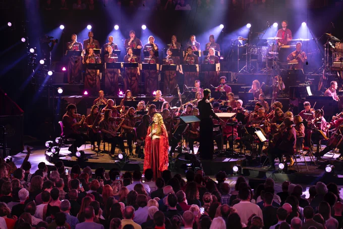 Florence Welch stands at the front of the stage, enveloped in a deep red gown, performing alongside the Jules Buckley Orchestra and choir at the Royal Albert Hall for Prom 69: Symphony of Lungs. The Jules Buckley Orchestra fills the stage, while a choir stands elevated in the background, illuminated by purple lights. Jules Buckley, dressed in black, directs the orchestra, while the audience watches from the Arena. 