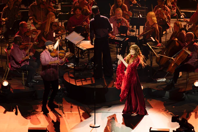A violinist from the Jules Buckley Orchestra wears a flat cap, standing to play his violin towards the front of the stage. On his left, Florence Welch claps her hands and smiles with glee, her red wavy hair flowing around her shoulders. Behind both of them, Jules Buckley conducts his orchestra – the front desks of the strings visible in the frame. 