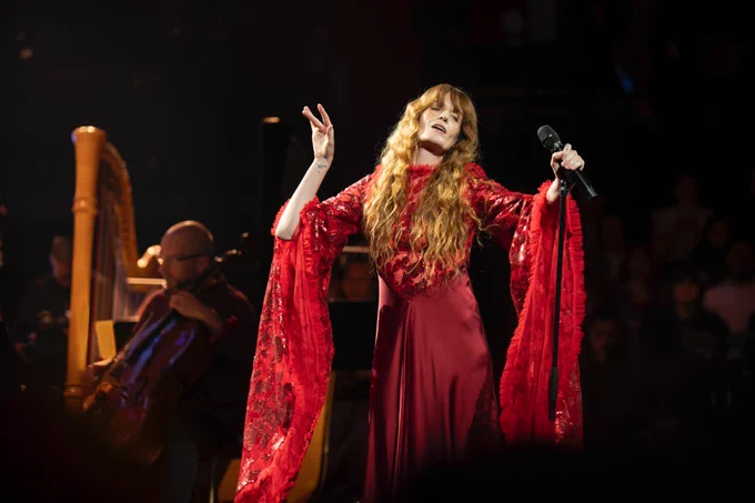 Florence Welch, lead singer of Florence   The Machine, performs passionately on stage at the Royal Albert Hall during Prom 69: Symphony of Lungs. She wears a flowing red gown with dramatic, lacy sleeves that extend as she lifts her arms. Her long, wavy red hair cascades down her shoulders. Behind her, musicians from the Jules Buckley Orchestra play string instruments, including a cello and a harp, under soft stage lighting.