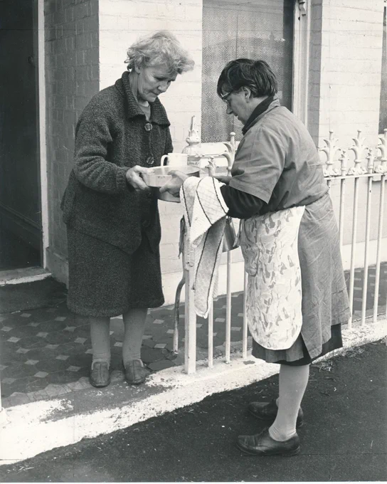 Two individuals exchanging a tray with tea cups outside a building.