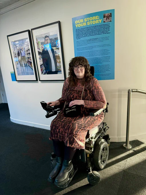 Shona, a white woman using a Powerchair, takes a photo in front of the start of her photography exhibition. There’s a blue sign introducing the exhibition and two of the photos. Shona is wearing an orange mesh dress, black boots and a black bag with a cats face on. She is smiling proudly.