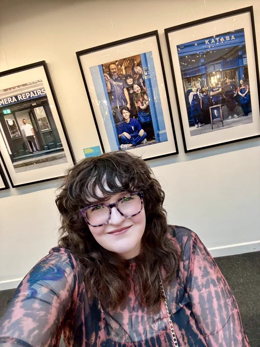 Shona, a white woman wearing glasses with a shag haircut, takes a selfie in front of some of the photos in her photography exhibition. She is smiling proudly.