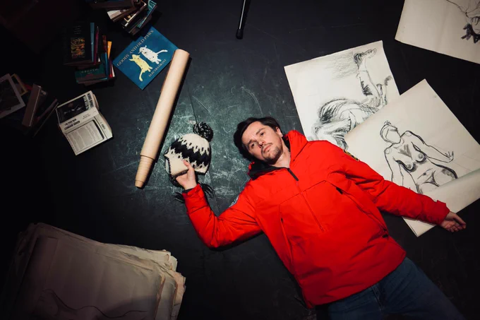 Promotional photo for A Version of Life: Man in red jacket lies on the ground atop books and sketches