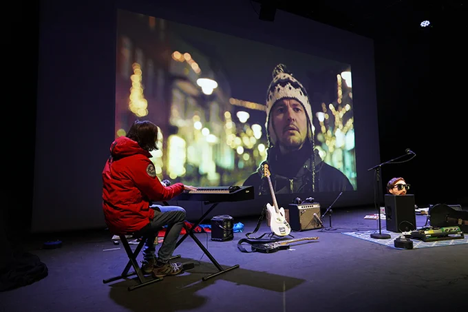 Promotional still for A Version of Life: Man in red jacket plays keyboard in front of a projected screen